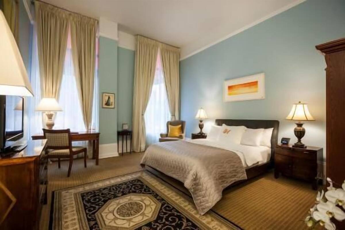 A hotel room with a bed, two bedside tables with lamps, a desk with a chair, a television, and a decorative rug.