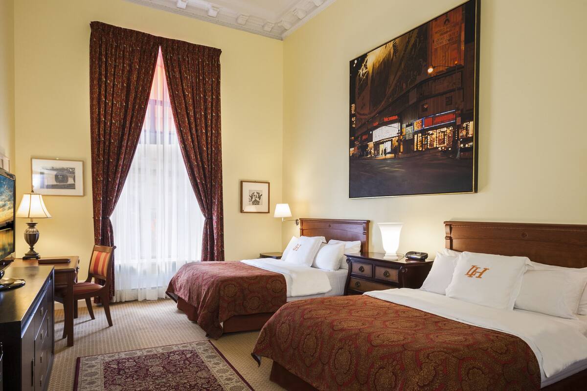 A hotel room with two beds, a desk, a chair, a large window with curtains, and a painting on the wall.