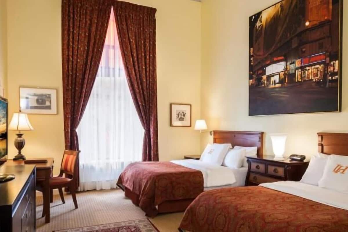 A hotel room with two beds, a desk, a chair, a lamp, and a large painting on the wall.