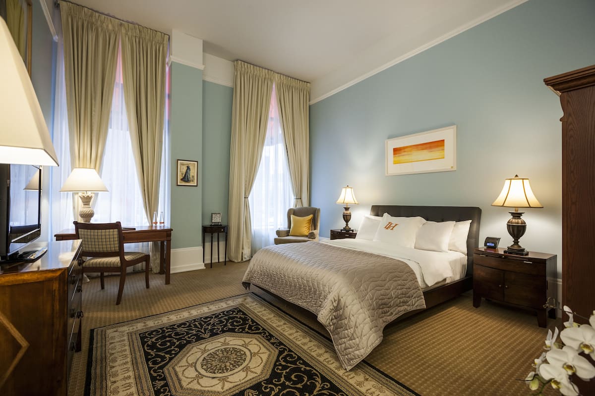 A hotel room with a large bed, two bedside tables with lamps, a desk with a chair, a television, and a decorative rug.