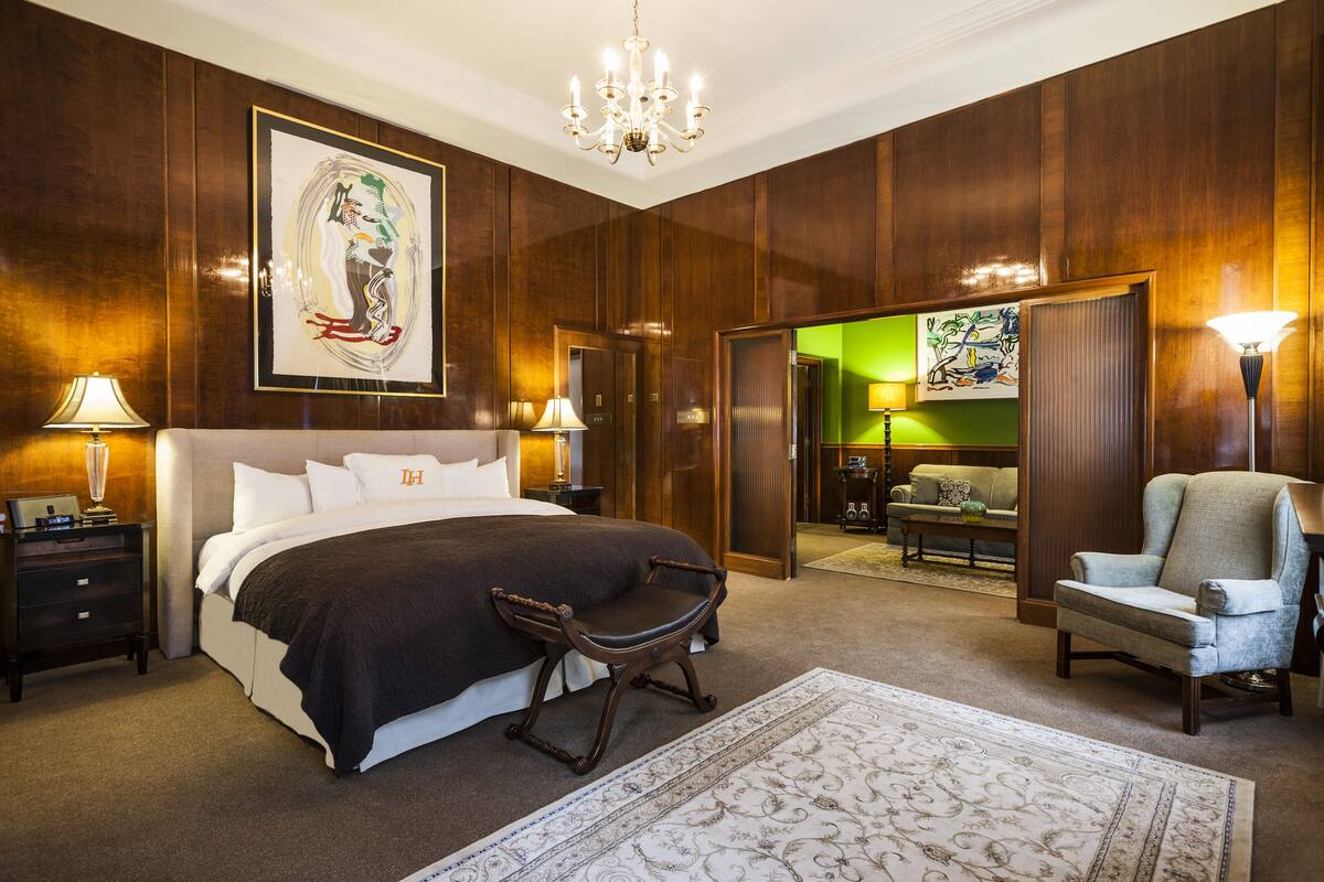 A spacious bedroom with a large bed, a sitting chair, a desk, and a chandelier.