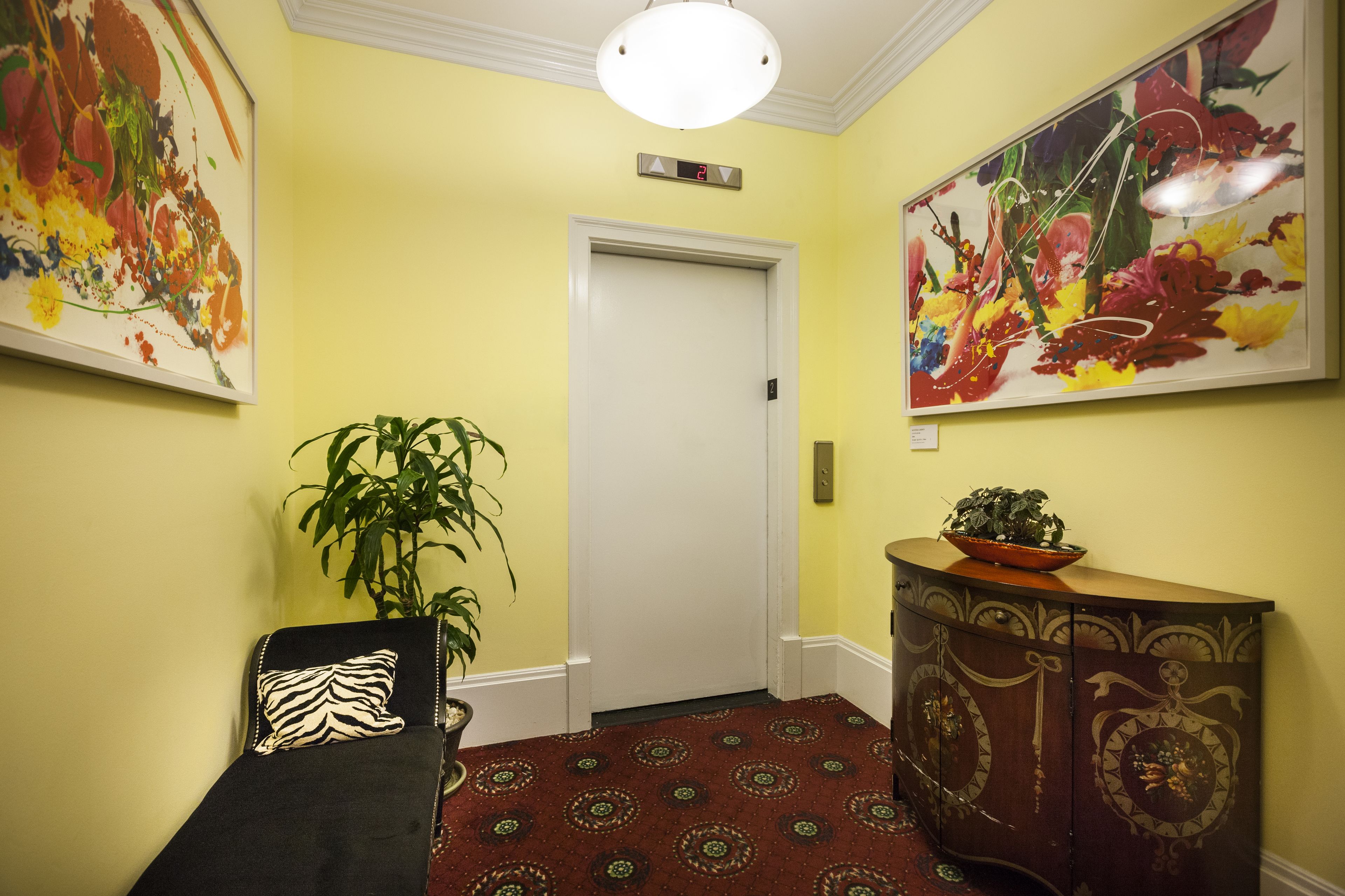 A hallway with yellow walls, a red patterned carpet, a black sofa with a zebra-patterned pillow, a potted plant, and a wooden cabinet with decorative carvings.