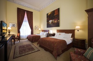 A hotel room with two beds, a desk, a chair, a TV, and a painting on the wall.
