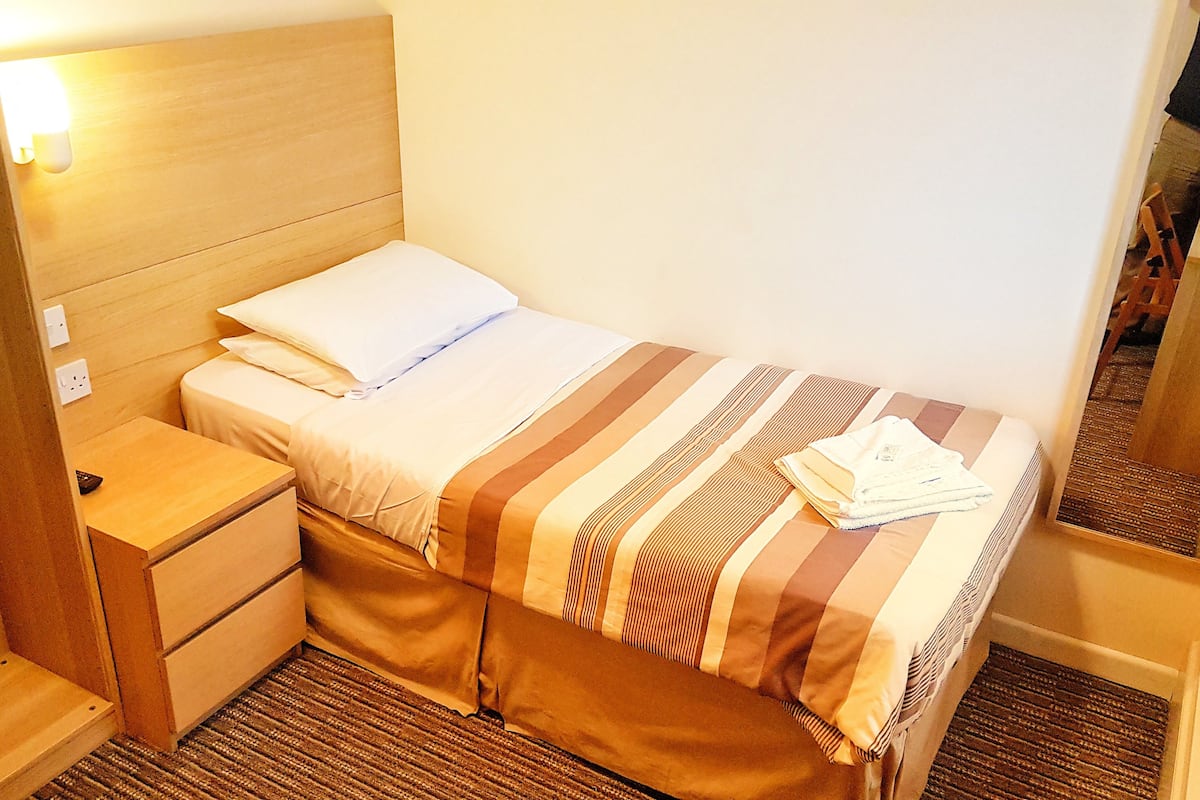 Una cama individual con una colcha a rayas, una mesita de noche de madera con cajón, y una lámpara fijada a la pared.