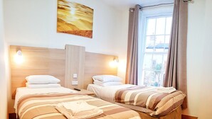 Una camera d'albergo con due letti, una grande finestra con tende, un quadro appeso al muro e una testiera in legno.