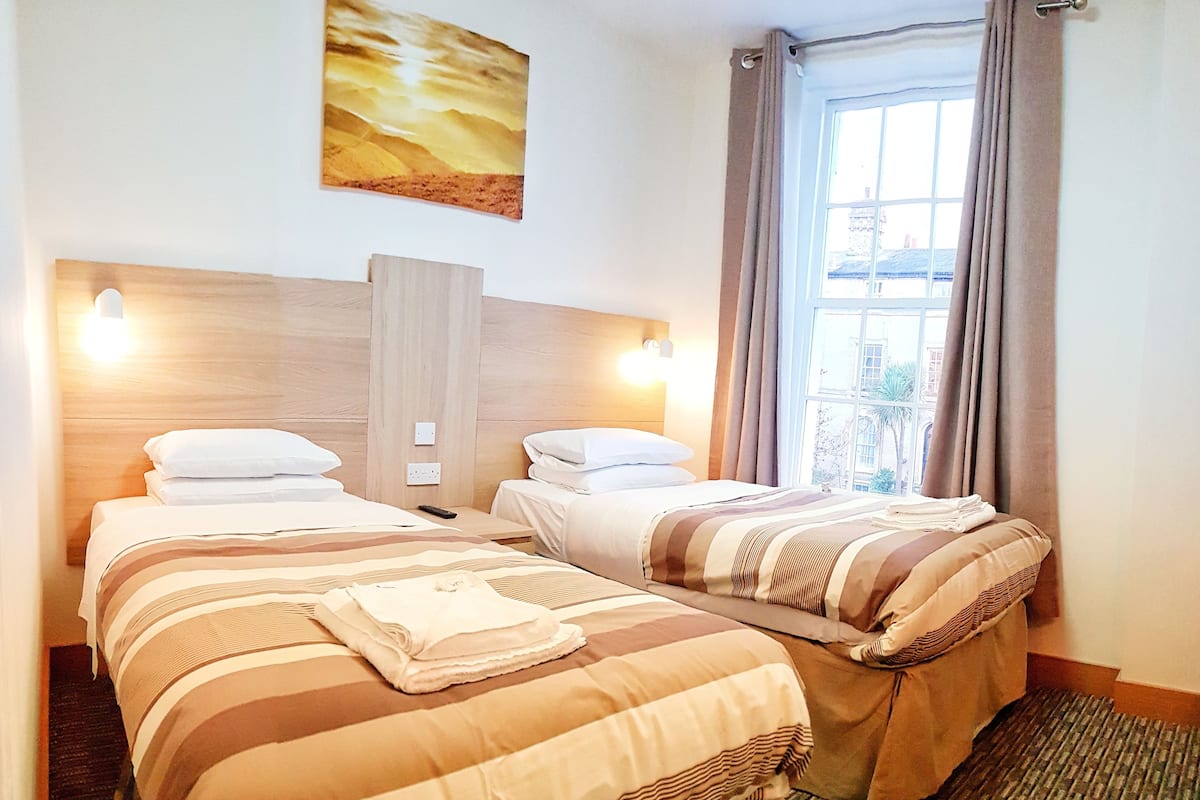 Habitación de hotel con dos camas, un ventanal con cortinas, un cuadro en la pared y cabecero de madera.