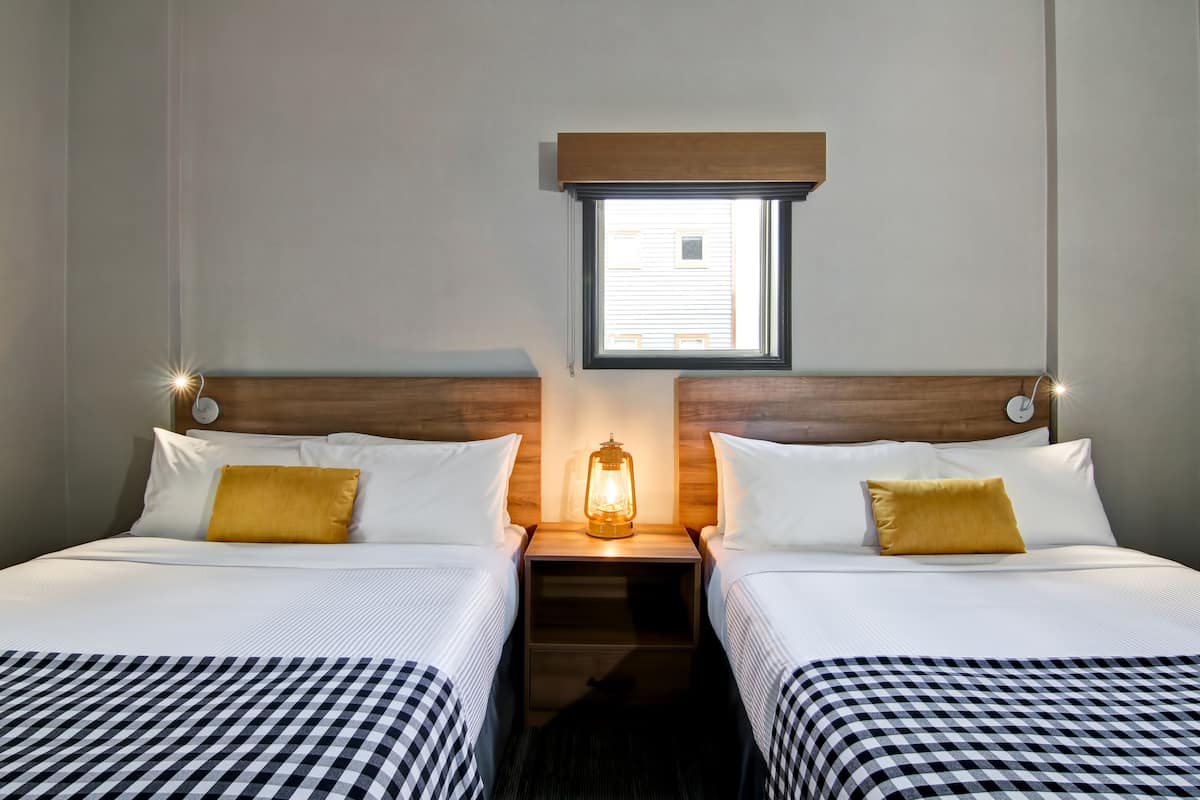 Quarto de hotel com duas camas, uma mesa de cabeceira com um candeeiro e uma janela, com um relógio na parede.
