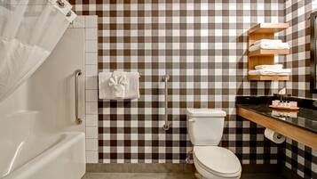 Une salle de bain avec un mur recouvert de carreaux à motifs, des toilettes blanches, une douche avec un rideau et une étagère en bois où sont posées des serviettes.