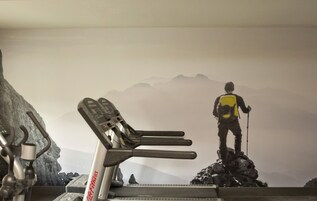 Una bicicleta estática de ejercicio marca Life Fitness, una persona de pie sobre una roca con una mochila y bastón de senderismo, y un paisaje montañoso de fondo.