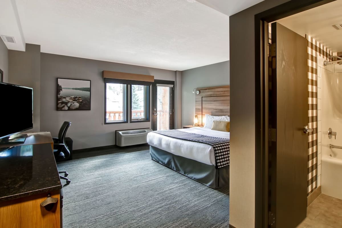 Quarto de hotel com cama, escrivaninha, cadeira, televisão, janela e porta.
