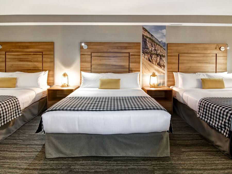 A hotel room with three beds, each with a wooden headboard, white bedding, and a checkered blanket. There are bedside lamps and a large window with a view of a mountainous landscape.