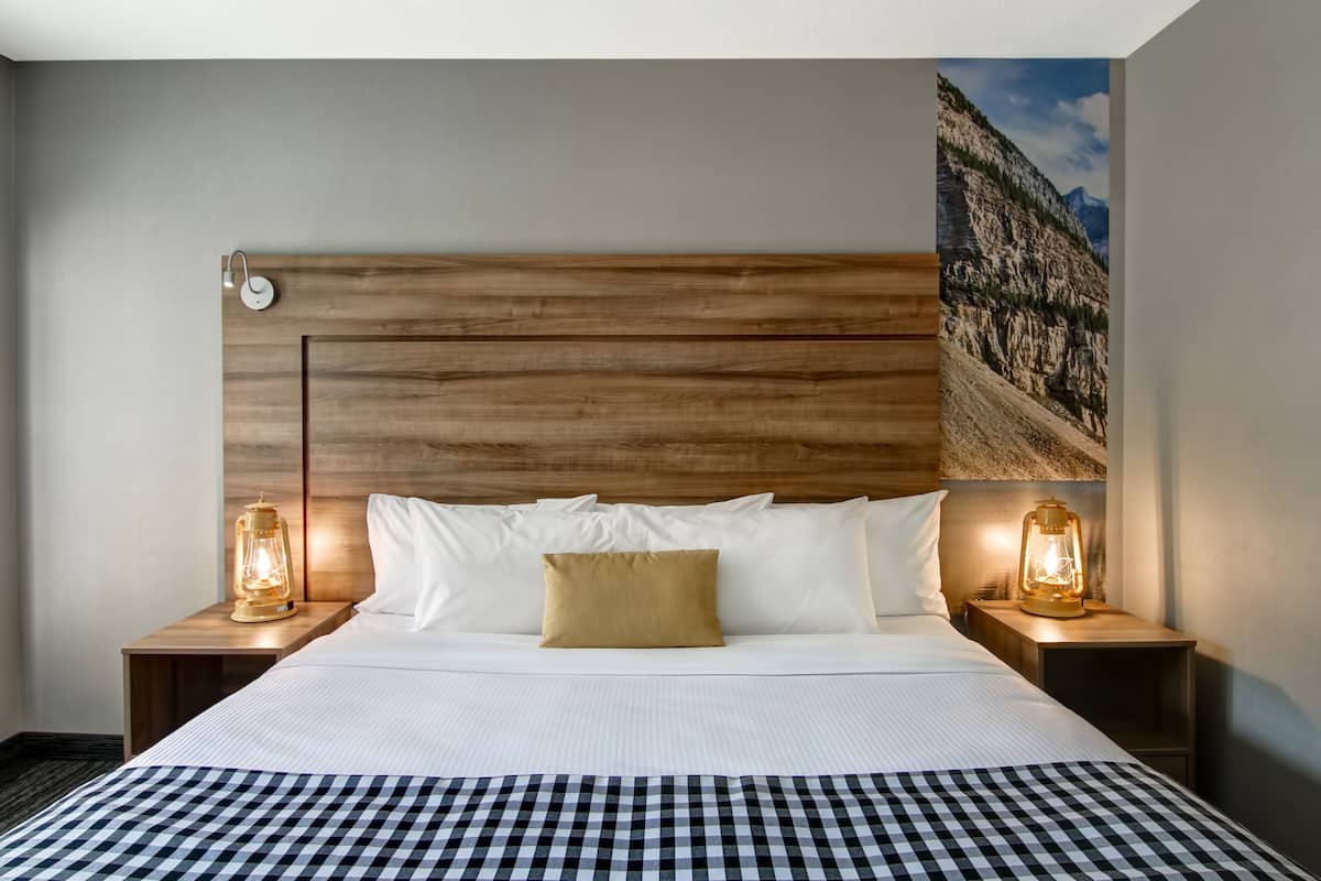 Quarto de hotel com cama grande, cabeceira de madeira, duas mesas de cabeceira com abajures em estilo de lanterna e uma pintura da paisagem de montanhas na parede.