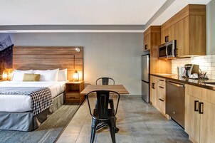 Una habitación de hotel moderna con una cama de cabecera de madera, una mesa de comedor con sillas, una zona de cocina con armarios de madera y electrodomésticos de acero inoxidable, y una mesita de noche con lámpara.