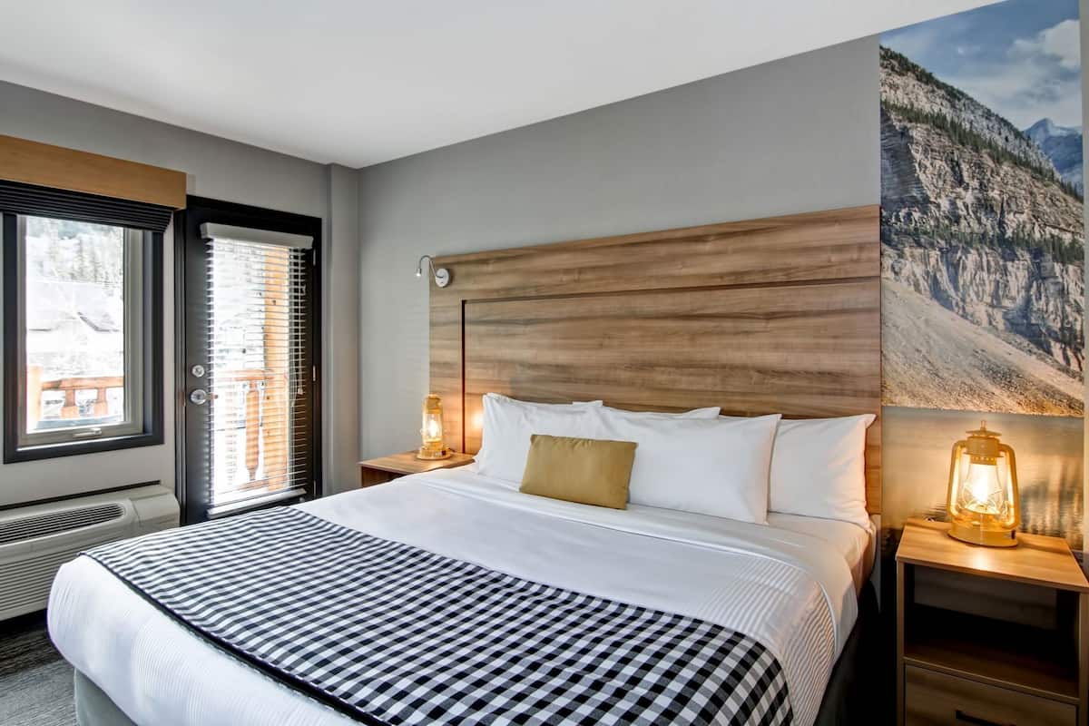 Quarto de hotel com cama grande, cabeceira de madeira, duas mesas de cabeceira com lanternas, janela com persianas e um mural com vista para as montanhas.