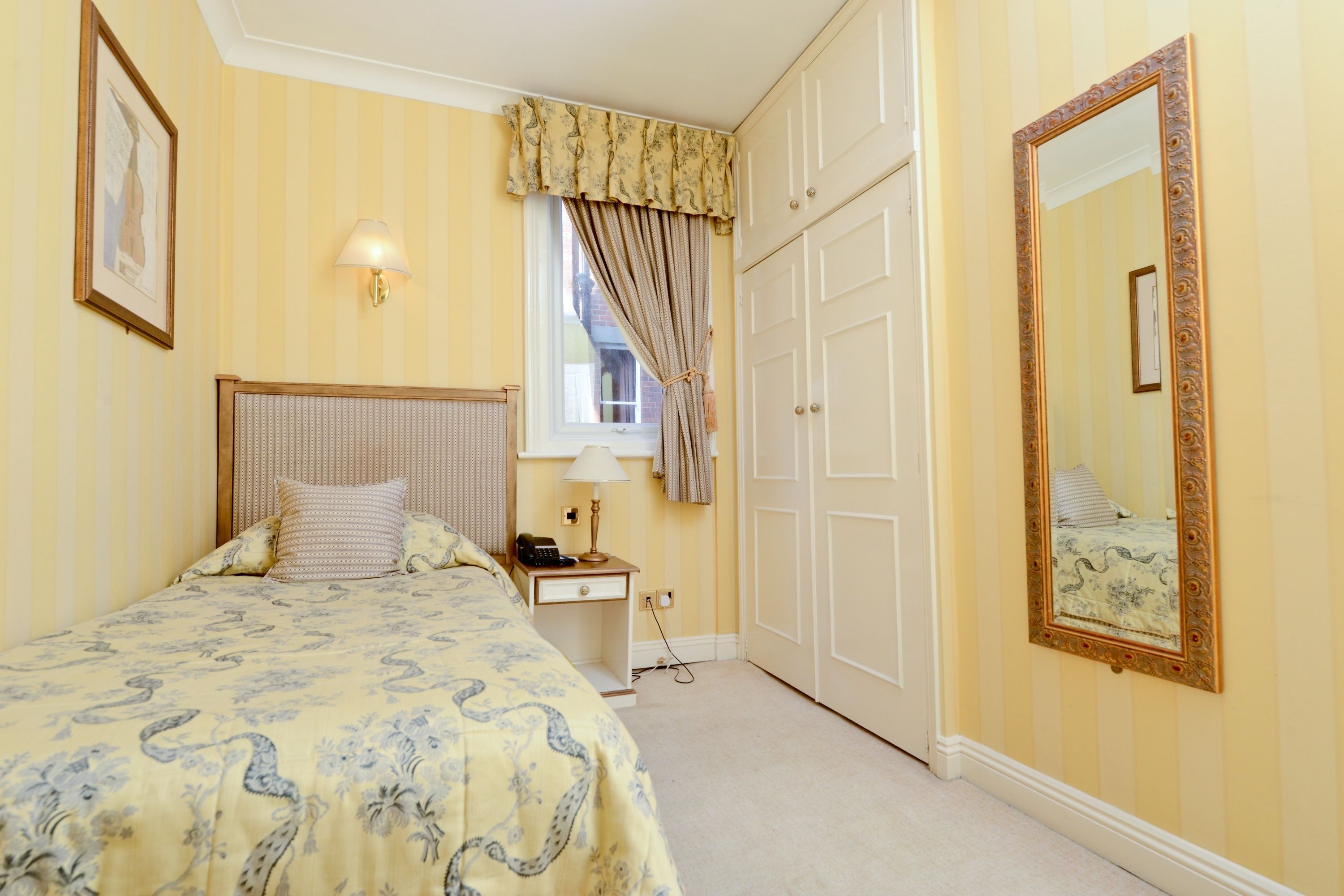 A bedroom with a bed, bedside table, lamp, window with curtains, wardrobe, and a mirror.