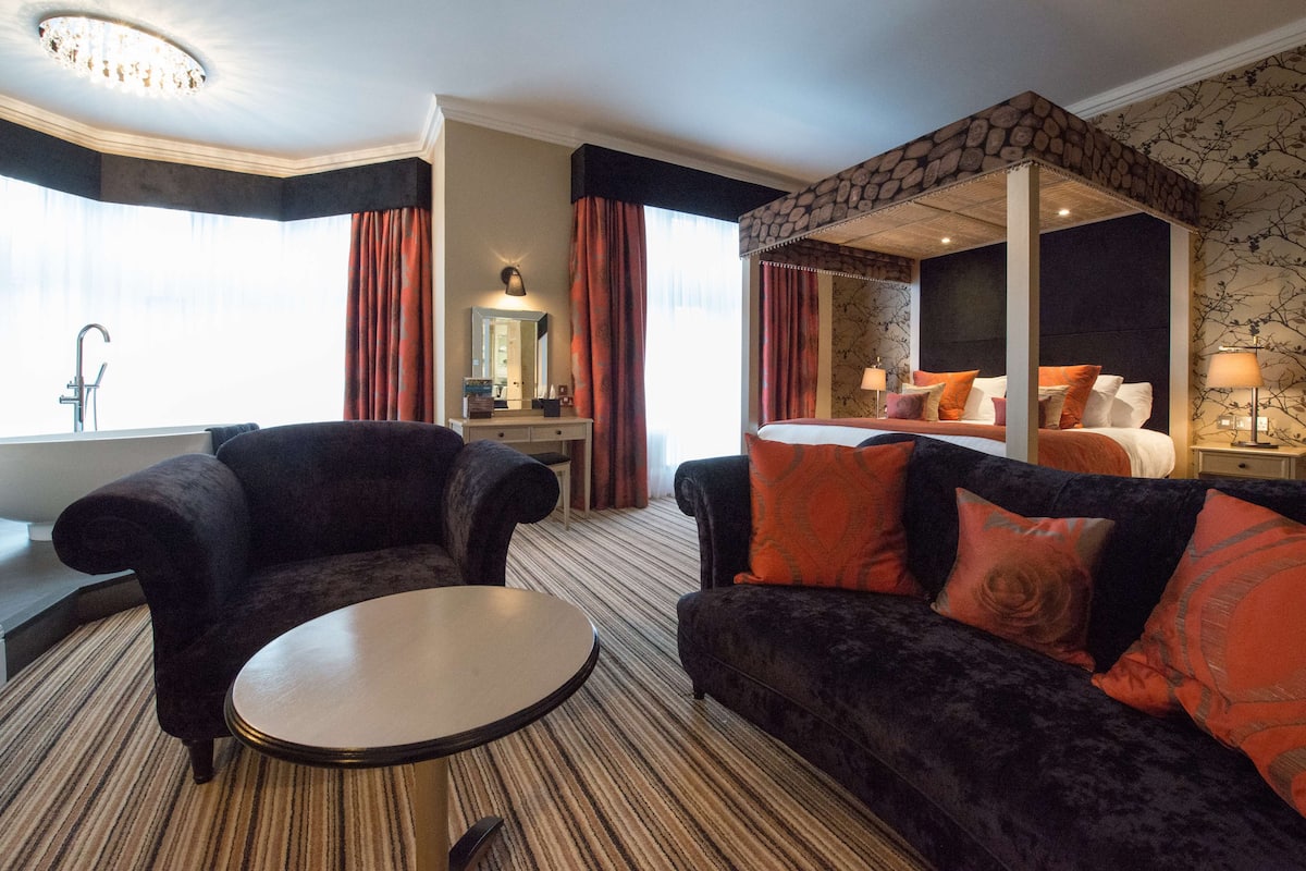 Ein Hotelzimmer mit einem großen Bett, einer Sitzecke mit rundem Tisch, einem Schminktisch mit Spiegel und einer Badewanne.