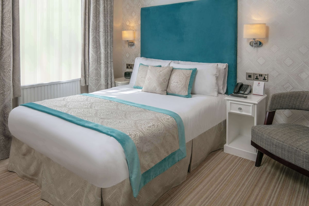 Ein ordentlich eingerichtetes Hotelzimmer mit einem großen Bett, einem Sessel, einem Nachttisch mit Lampe und einem Telefon.
