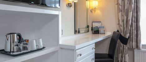 A hotel room with a white dresser, a black chair, a tray with a kettle and cups, a flat-screen TV, and a mirror with wall sconces.