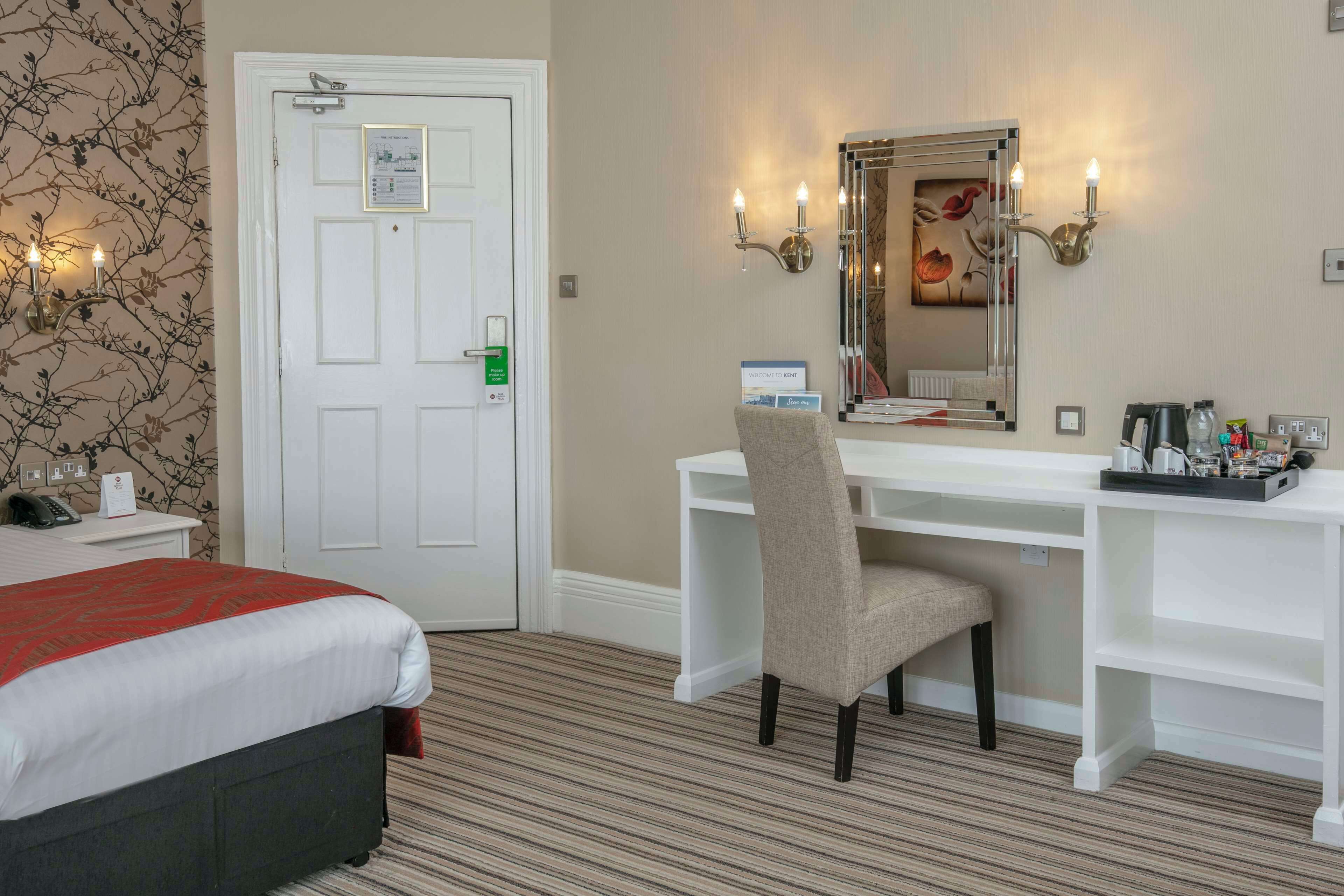 A hotel room with a bed, a desk with a chair, a mirror, and a wall-mounted light fixture.