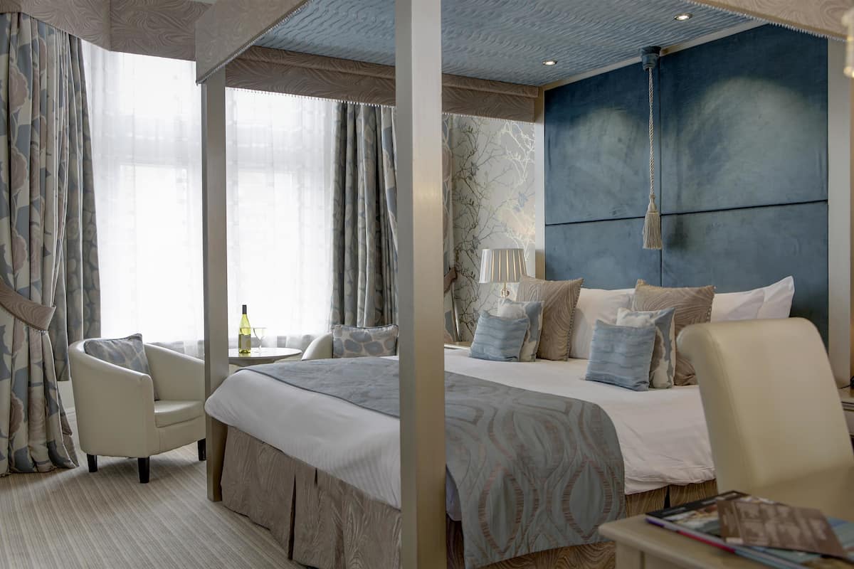 Ein Hotelzimmer mit einem großen Bett, zwei Sesseln, einem Nachttisch und Blick auf die Stadt durch große Fenster.