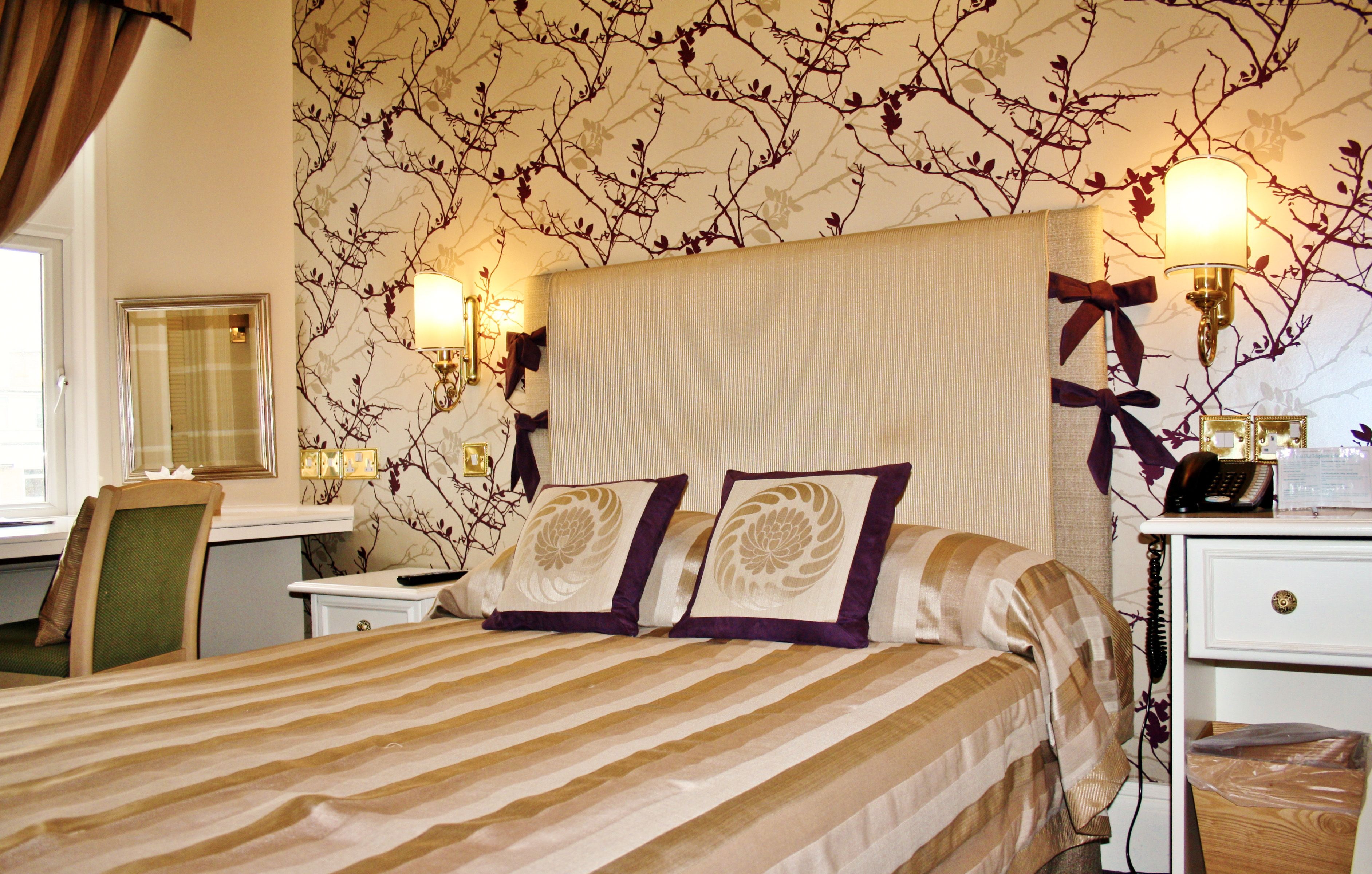 Ein Hotelzimmer mit Bett, Schreibtisch, Stuhl, Nachttisch und einer Wand mit floralem Wandpapier.