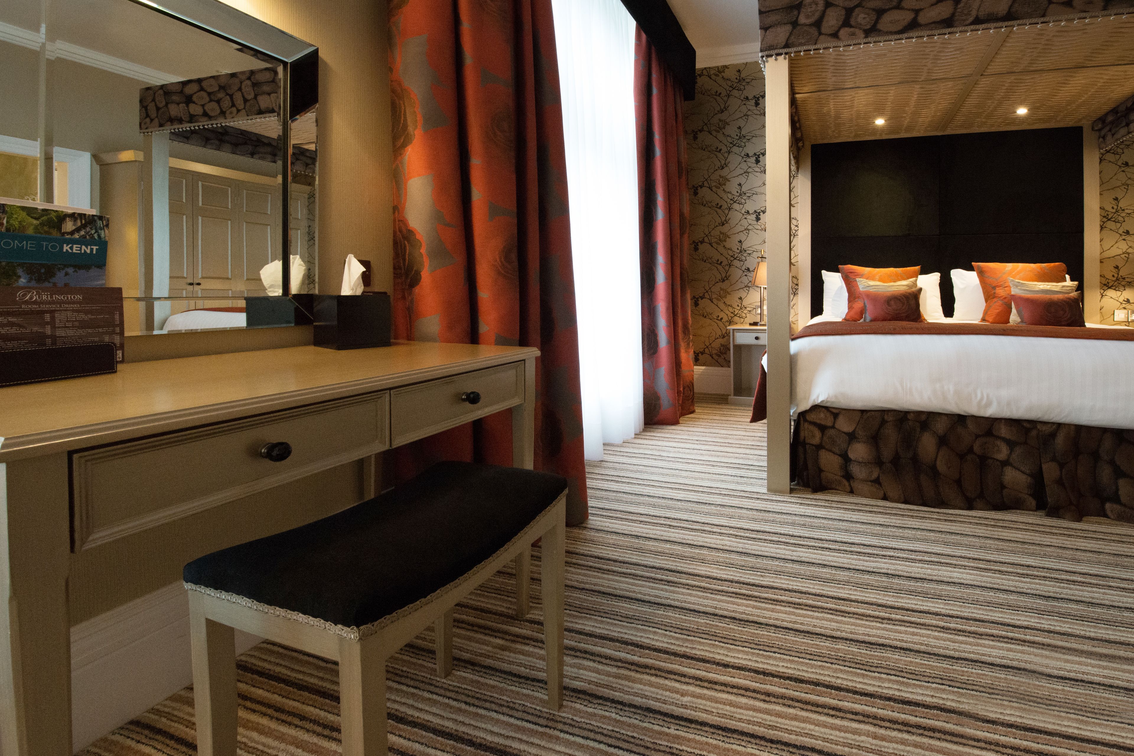 A hotel room with a bed, a desk, a bench, a mirror, and curtains.