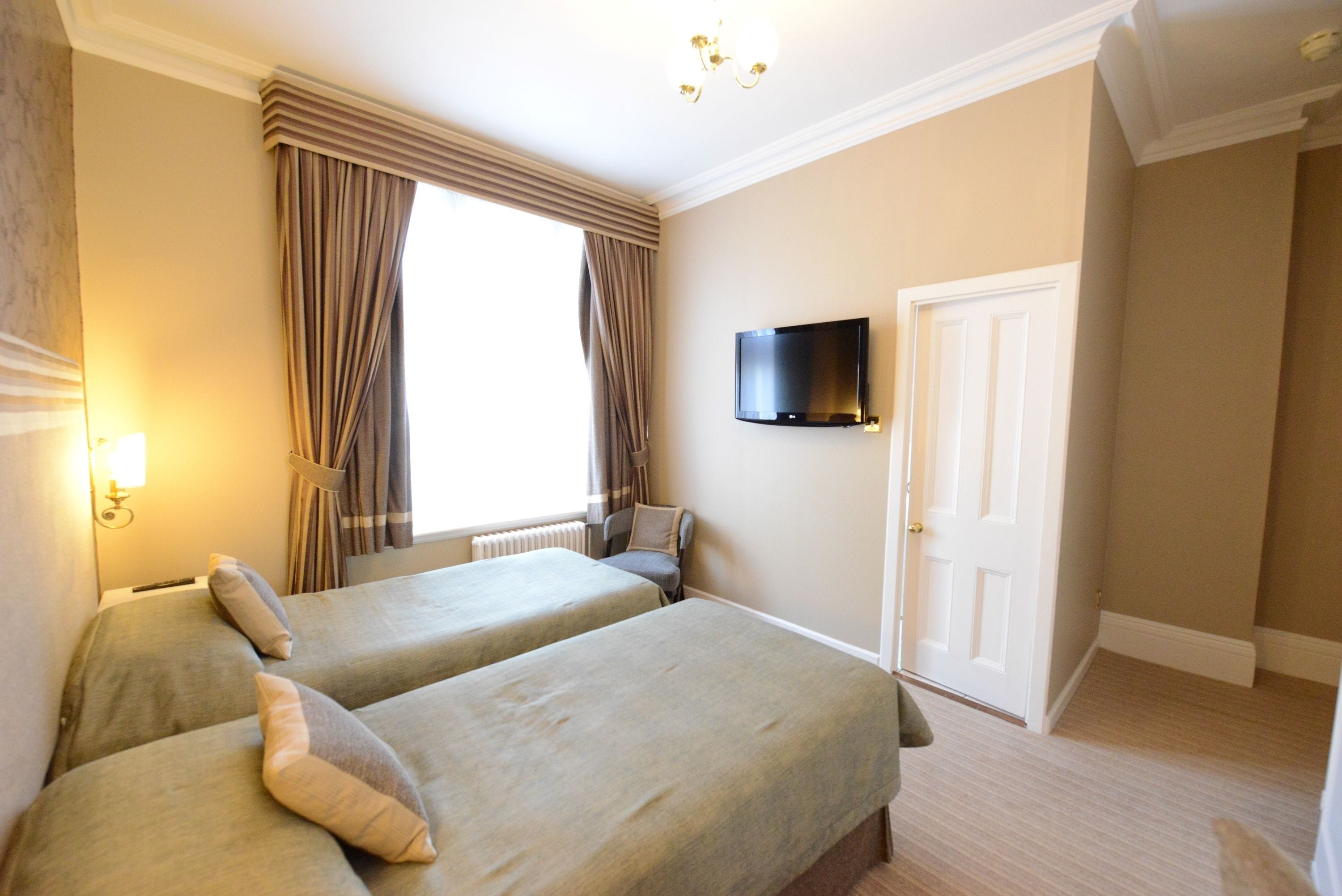 A hotel room with two beds, a television on the wall, a window with curtains, and a door.