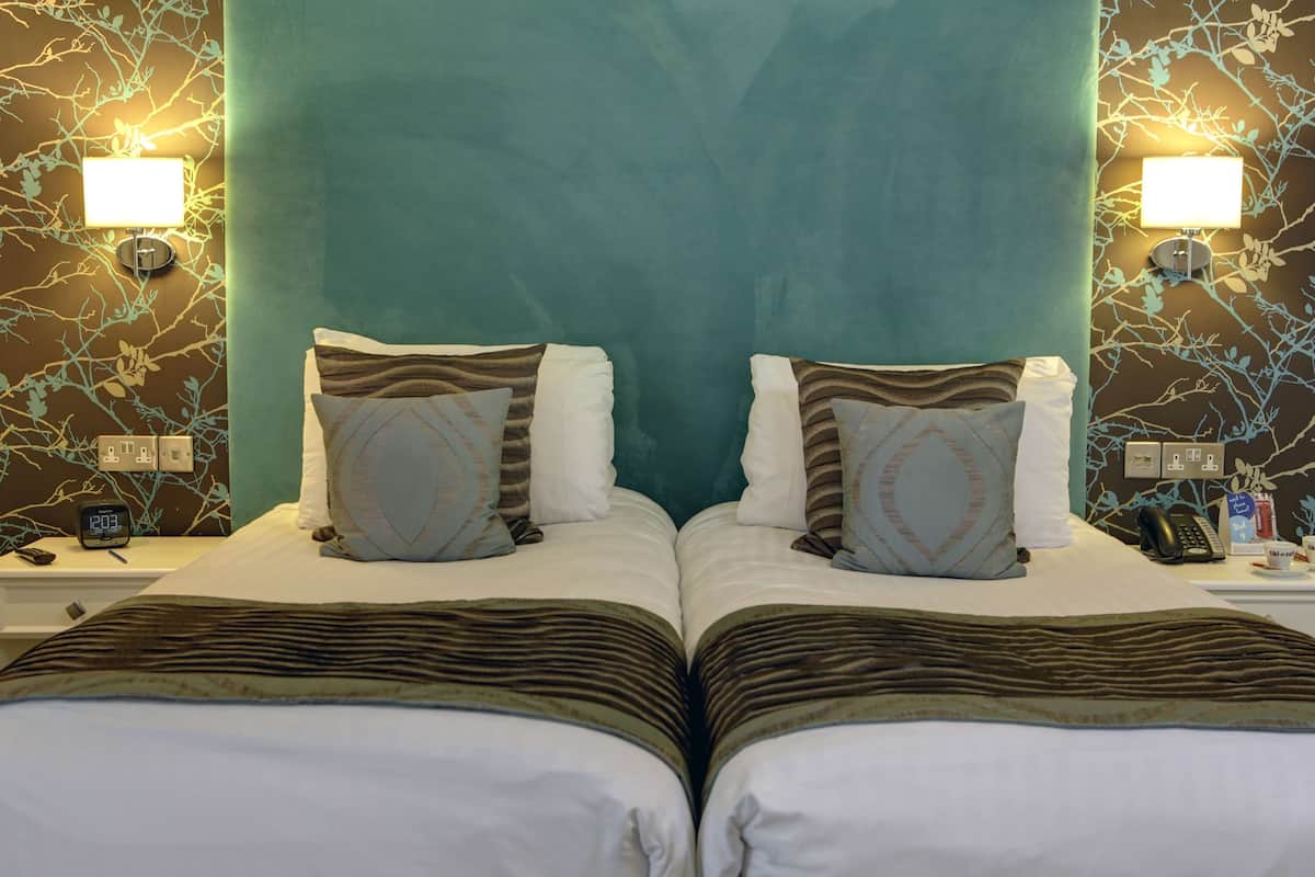 Ein Hotelzimmer mit zwei Betten, einem türkisblauen Kopfteil, gemusterten Kissen, Nachttischen mit Lampen und einer Uhr.