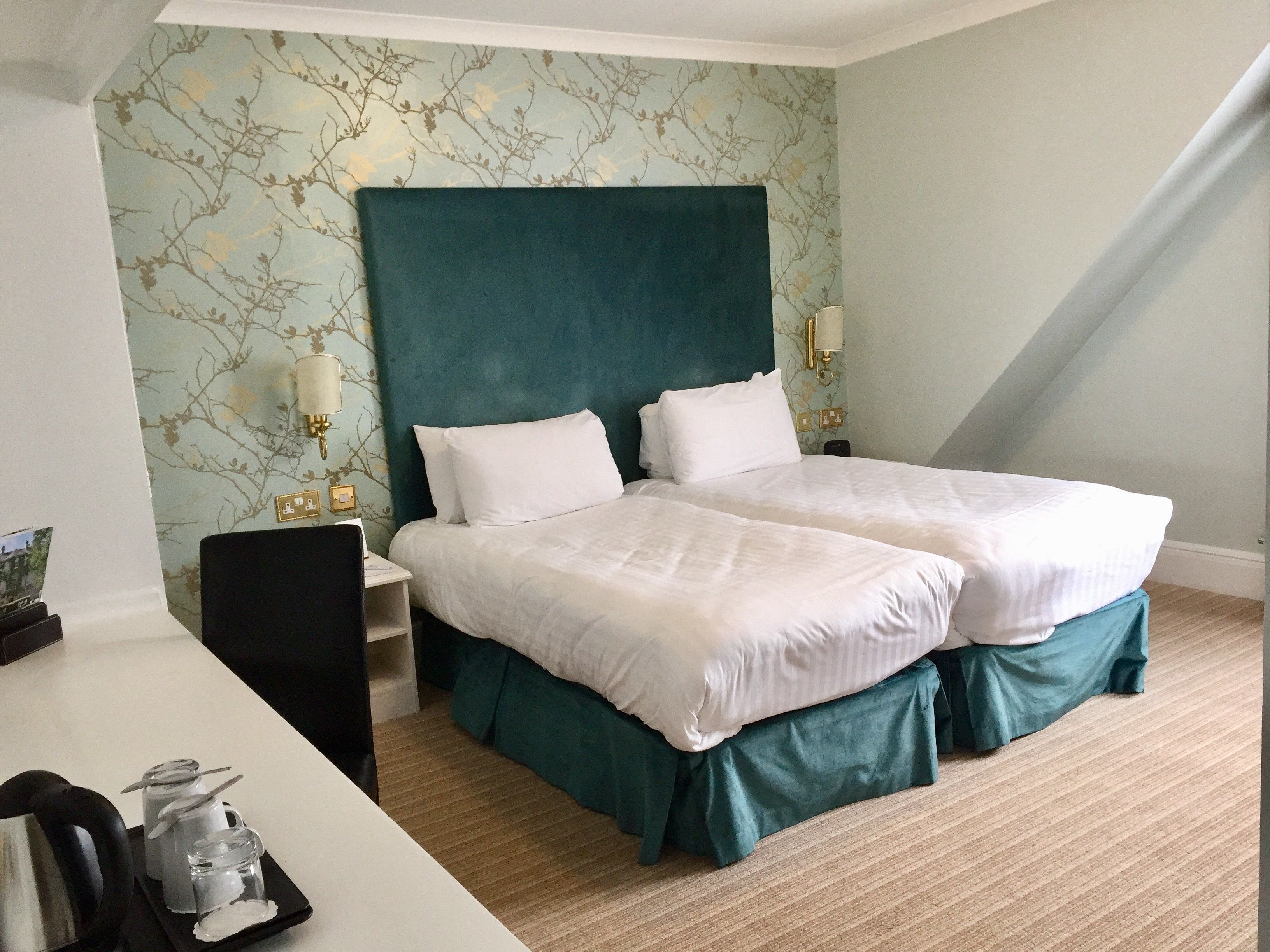 A hotel room with two beds, a desk, and a chair. There is a small table with a pitcher and glasses. The walls have a floral wallpaper pattern.