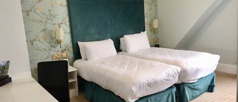 A hotel room with two beds, a desk, and a chair. There is a small table with a pitcher and glasses. The walls have a floral wallpaper pattern.