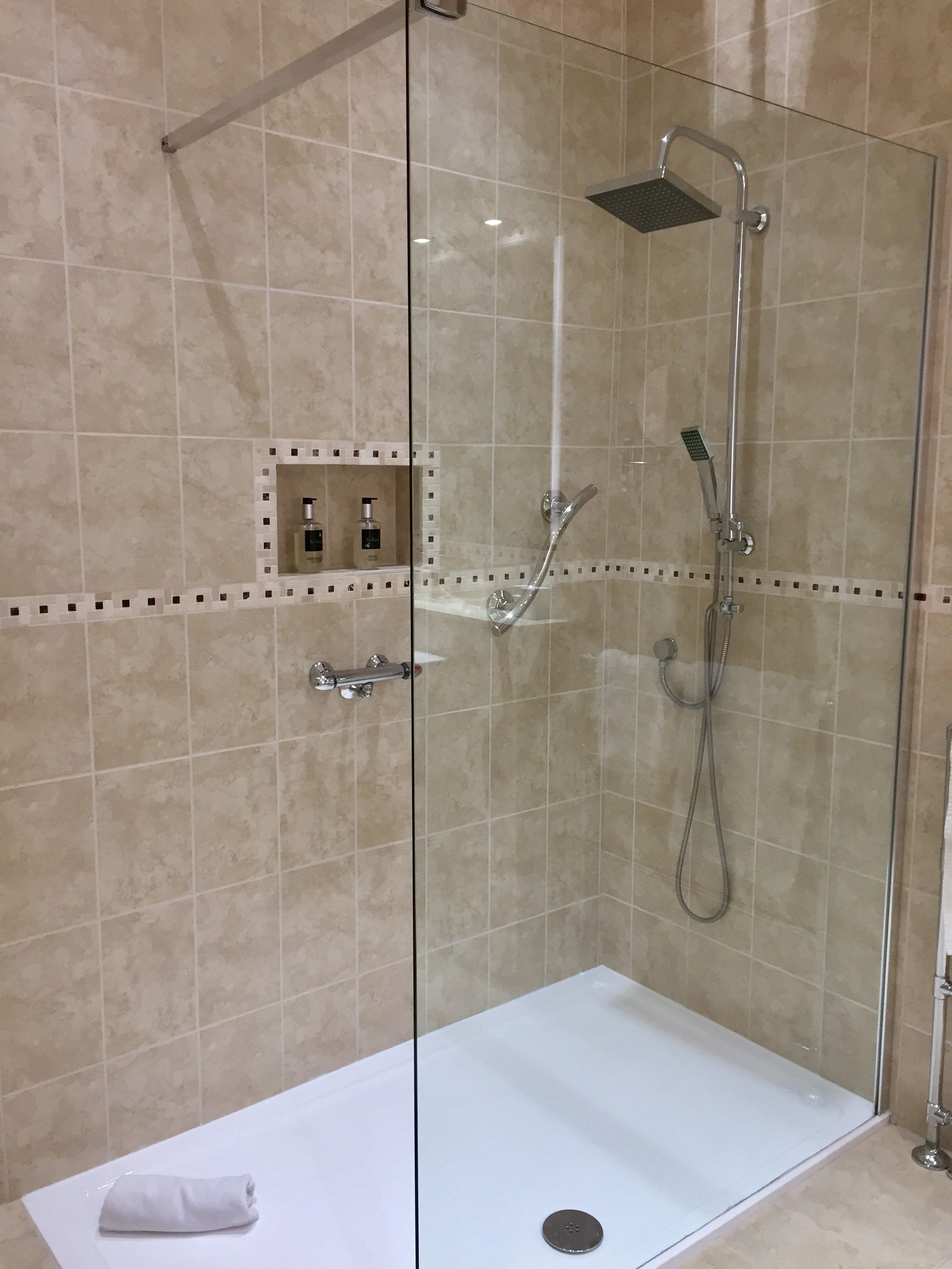 A glass-enclosed shower area with a handheld showerhead, a wall-mounted faucet, and a handheld showerhead. There is a small shelf with toiletries.