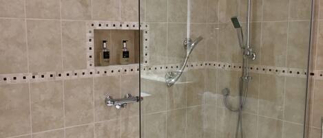 A glass-enclosed shower area with a handheld showerhead, a wall-mounted faucet, and a handheld showerhead. There is a small shelf with toiletries.
