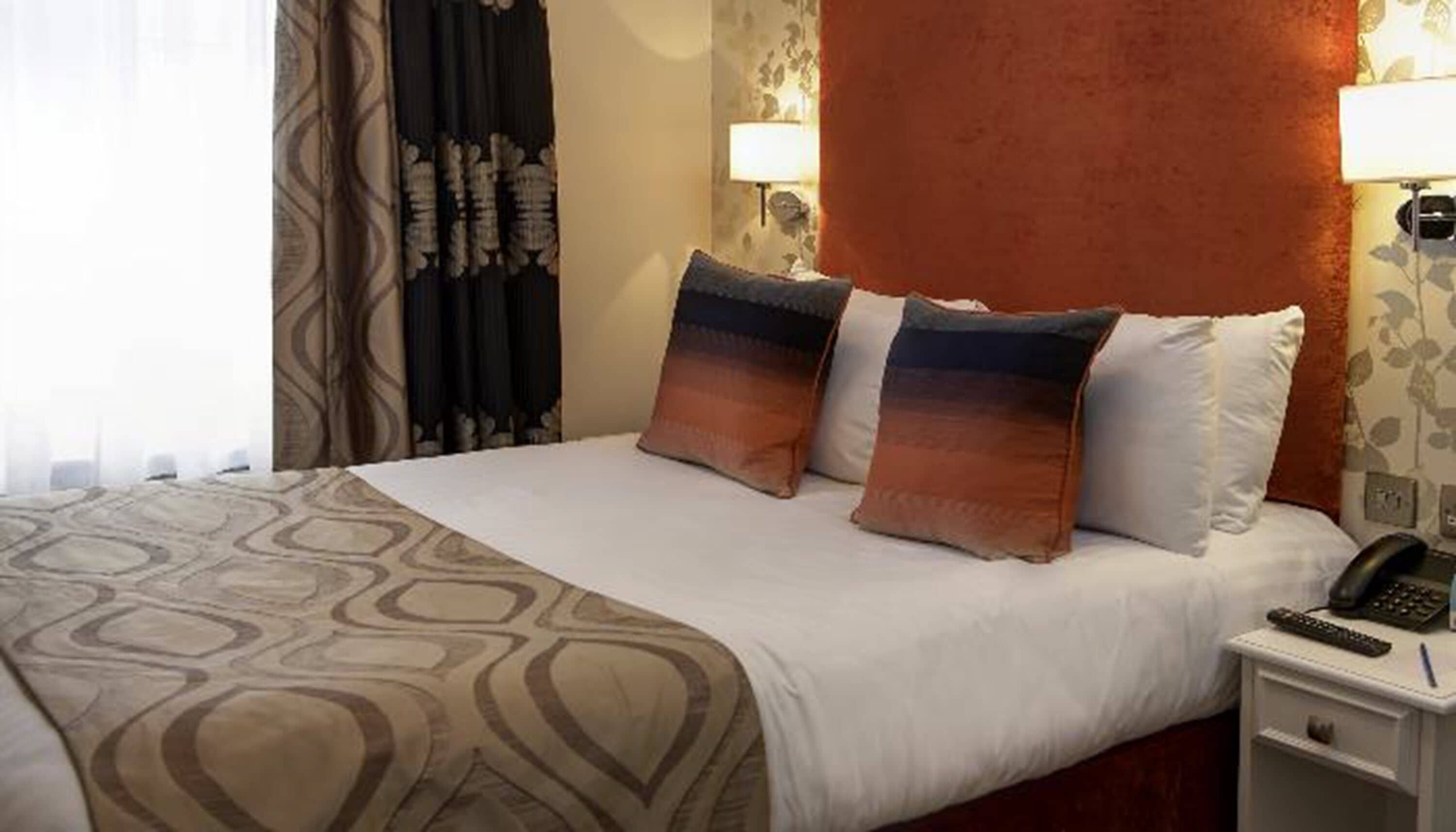 Una habitación de hotel con una cama, dos almohadas, una mesita de noche con un teléfono y una lámpara.