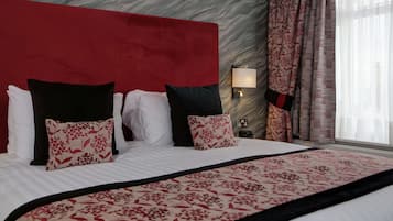 A hotel room with a large bed, red headboard, floral-patterned pillows, a nightstand with a lamp, and curtains.