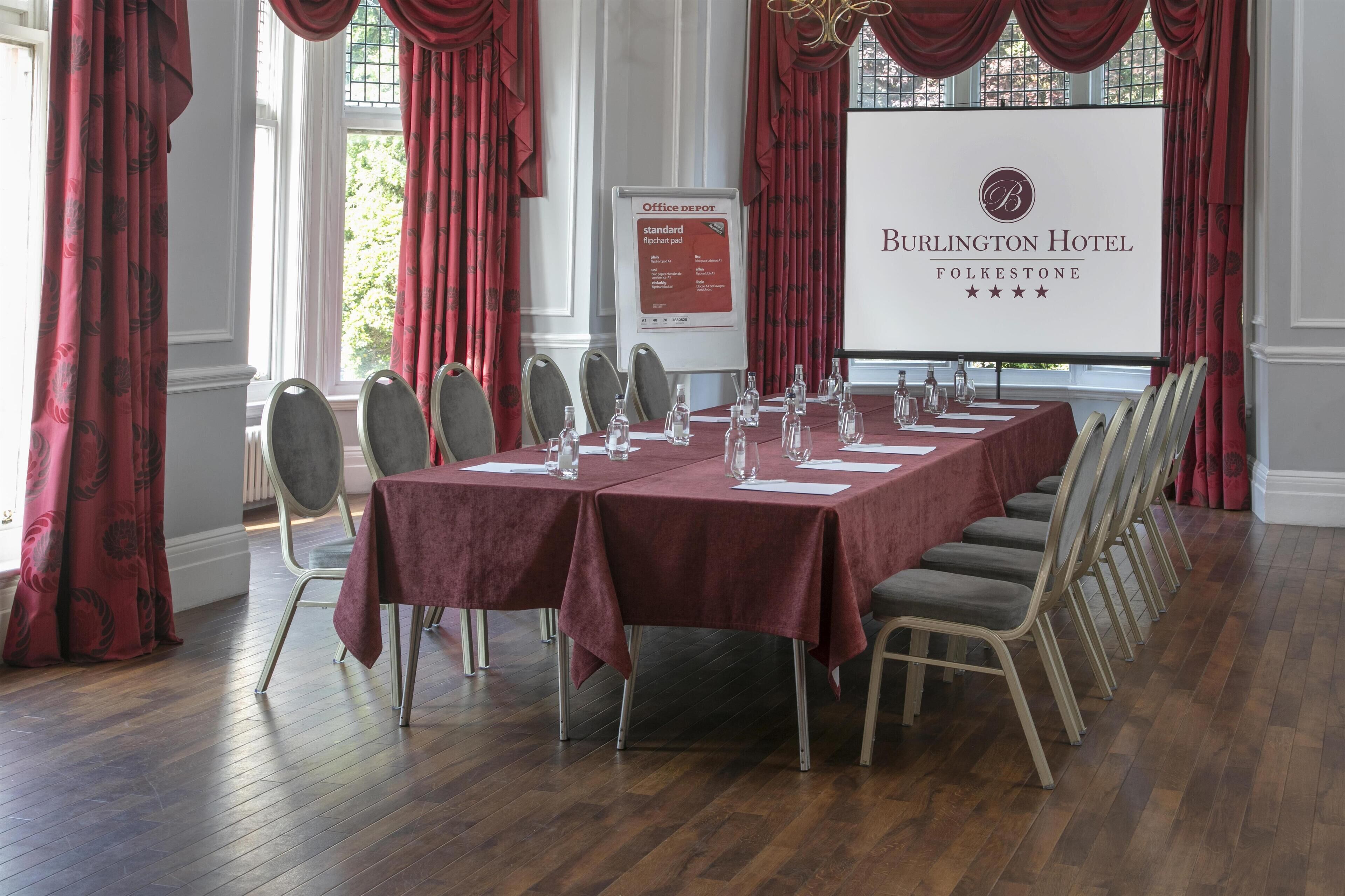 A conference room at Burlington Hotel with a long table, chairs, and a projector screen displaying the hotel name and amenities.