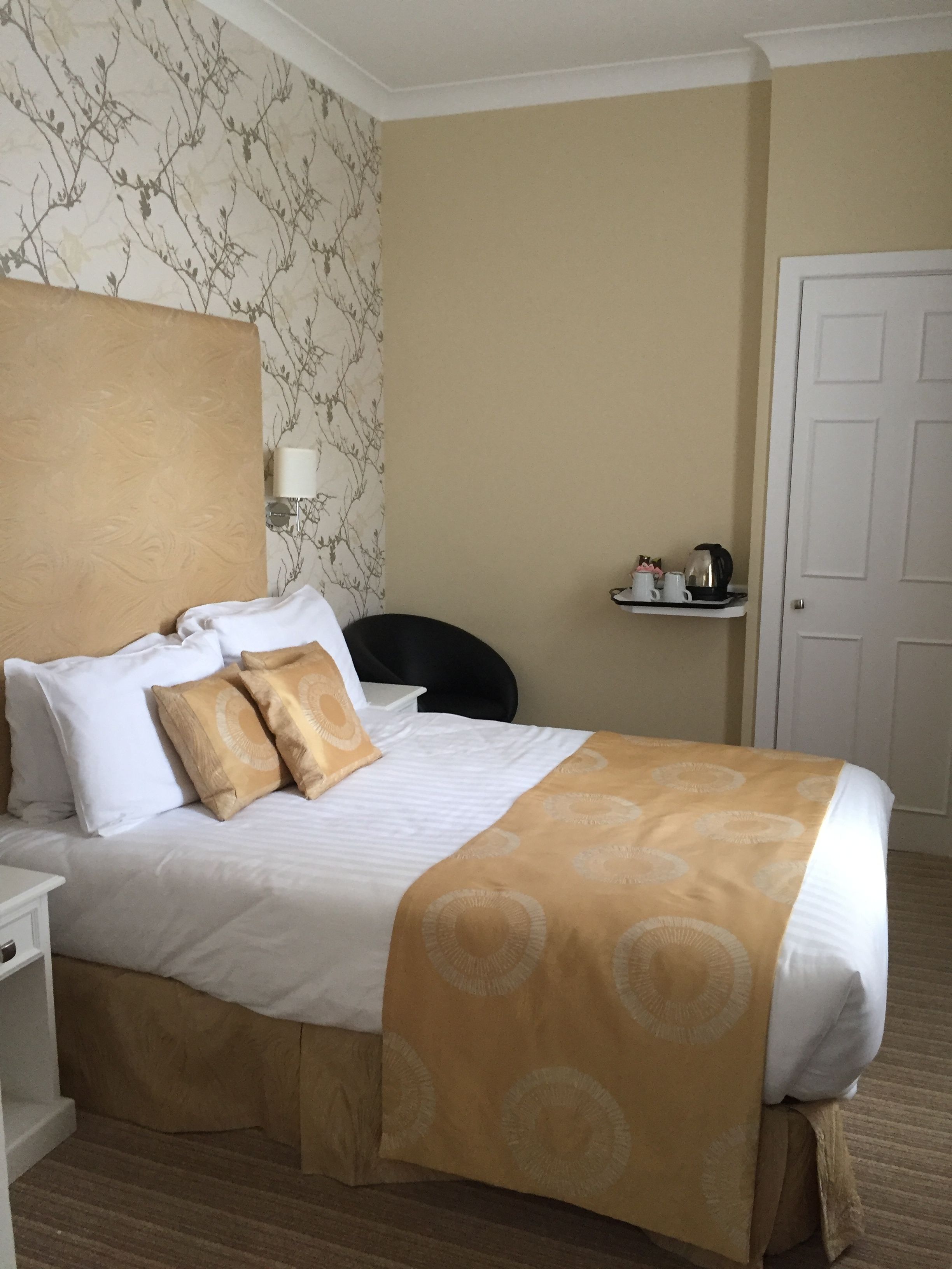 Una cama bien hecha con una colcha blanca y beige, dos almohadas blancas y dos cojines decorativos beige. Hay una mesita de noche blanca con una lámpara, una silla negra y un pequeño estante con una cafetera y tazas. La habitación tiene una pared beige con un estampado floral de papel tapiz y una puerta blanca.