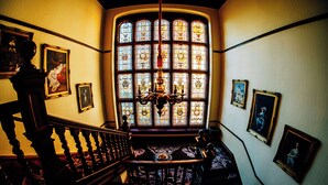 Uma escadaria imponente com uma janela de vitrais acima, obras de arte emolduradas nas paredes e um lustre.