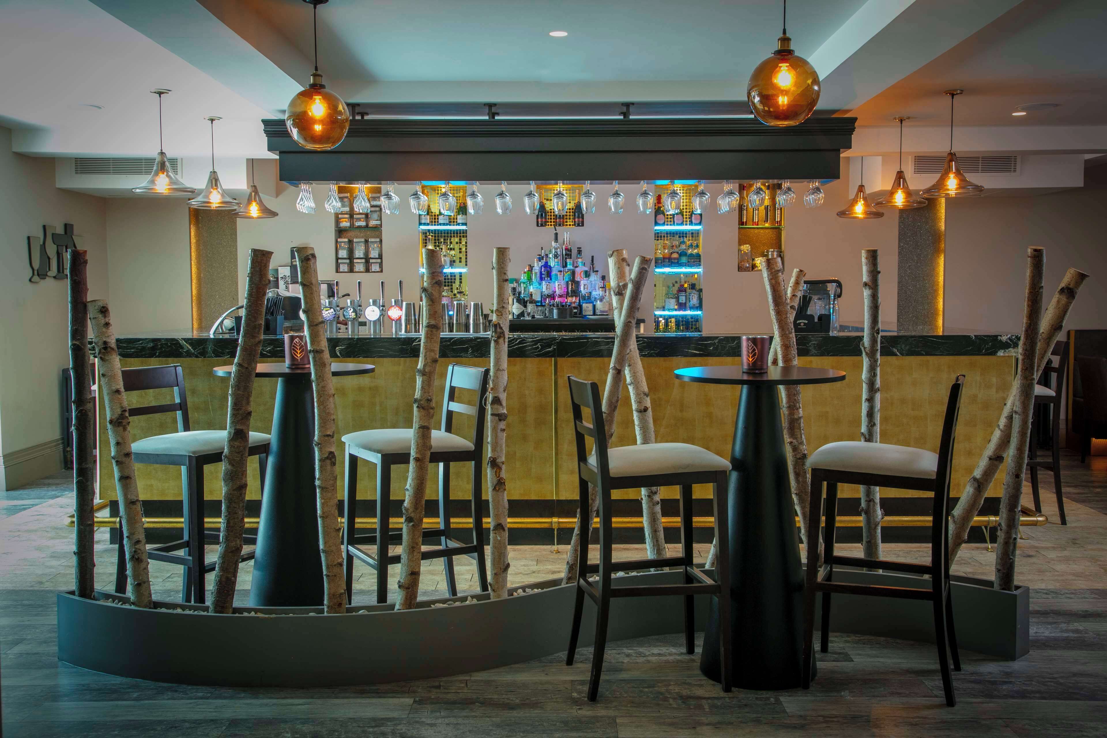 A bar area with a modern design, featuring a bar counter, hanging light fixtures, and a seating arrangement with chairs and tables.