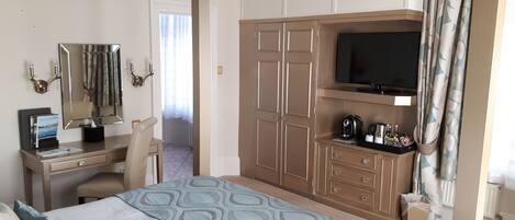 Habitación de hotel con cama, escritorio y silla, armario, televisor y ventana con cortinas.