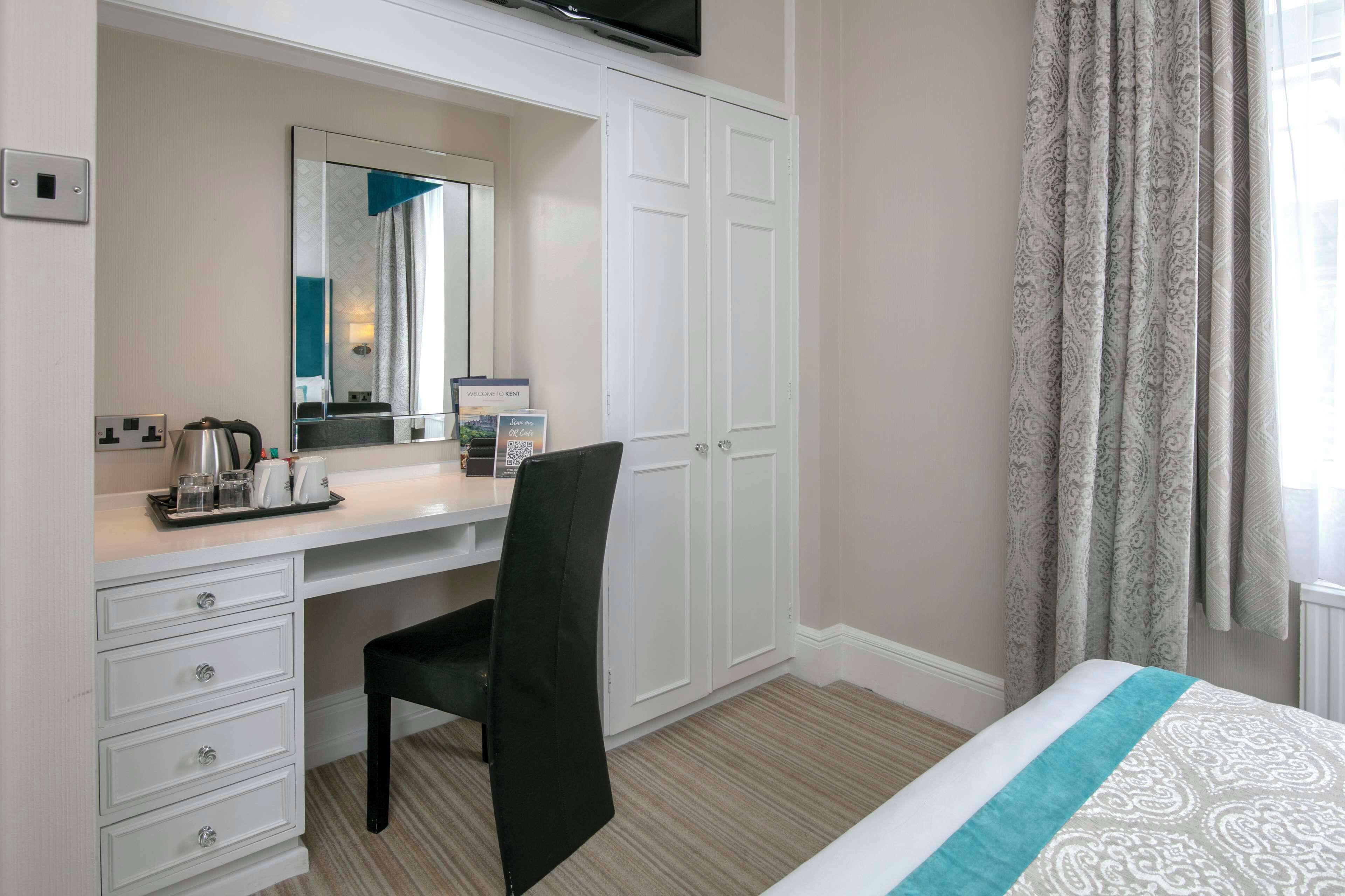 Une chambre d’hôtel avec un bureau blanc, une chaise noire, un lit recouvert d’une couvre-lit à motifs, un grand miroir et une armoire.