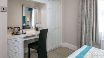 Une chambre d’hôtel avec un bureau blanc, une chaise noire, un lit recouvert d’une couvre-lit à motifs, un grand miroir et une armoire.
