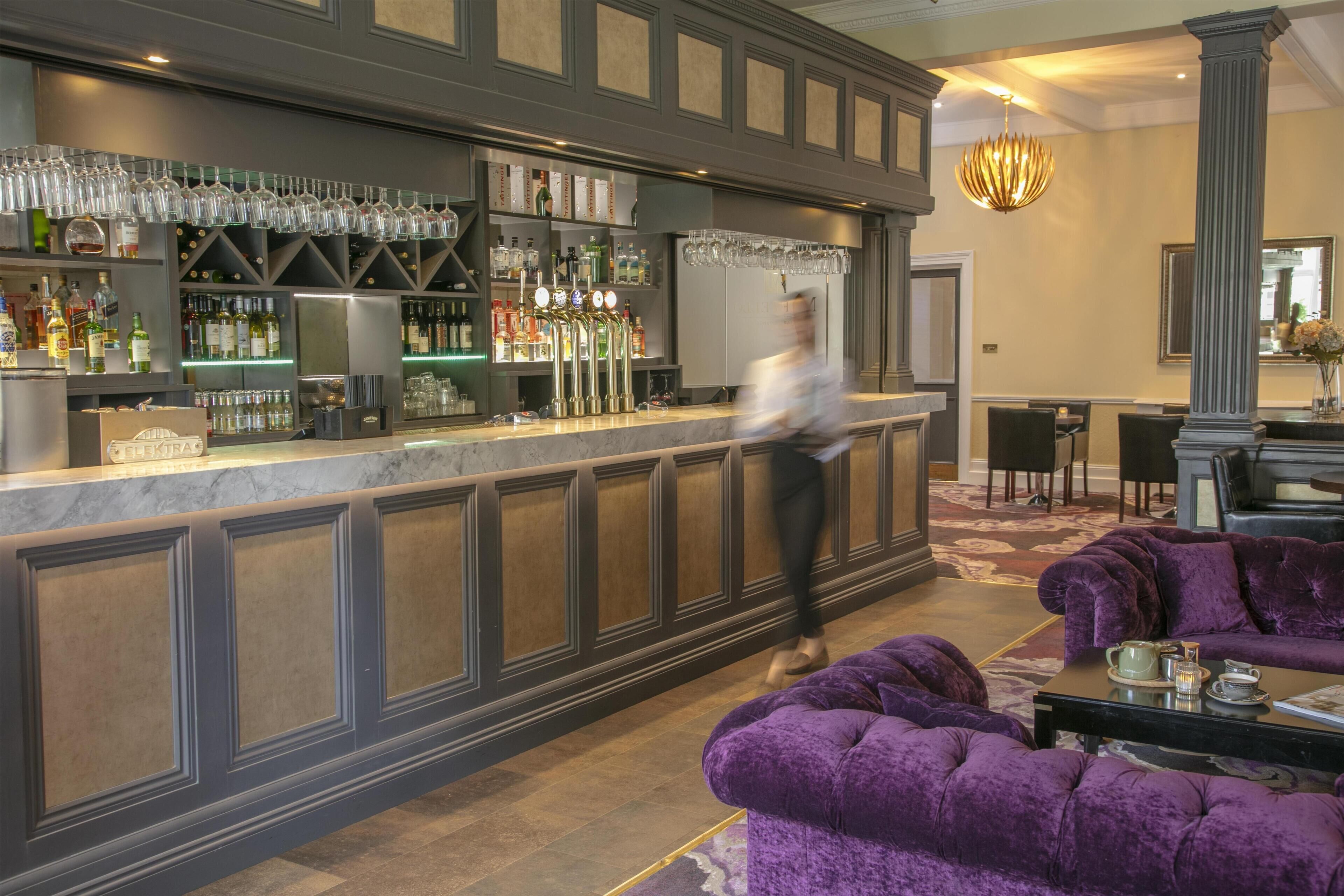 A bar area with a marble countertop, shelves with bottles, and a bartender. There is a seating area with a purple sofa and a table with a teapot.
