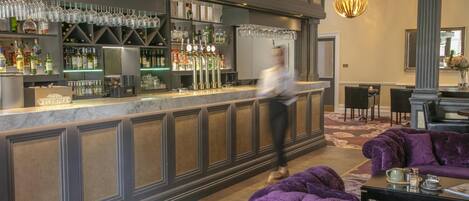A bar area with a marble countertop, shelves with bottles, and a bartender. There is a seating area with a purple sofa and a table with a teapot.