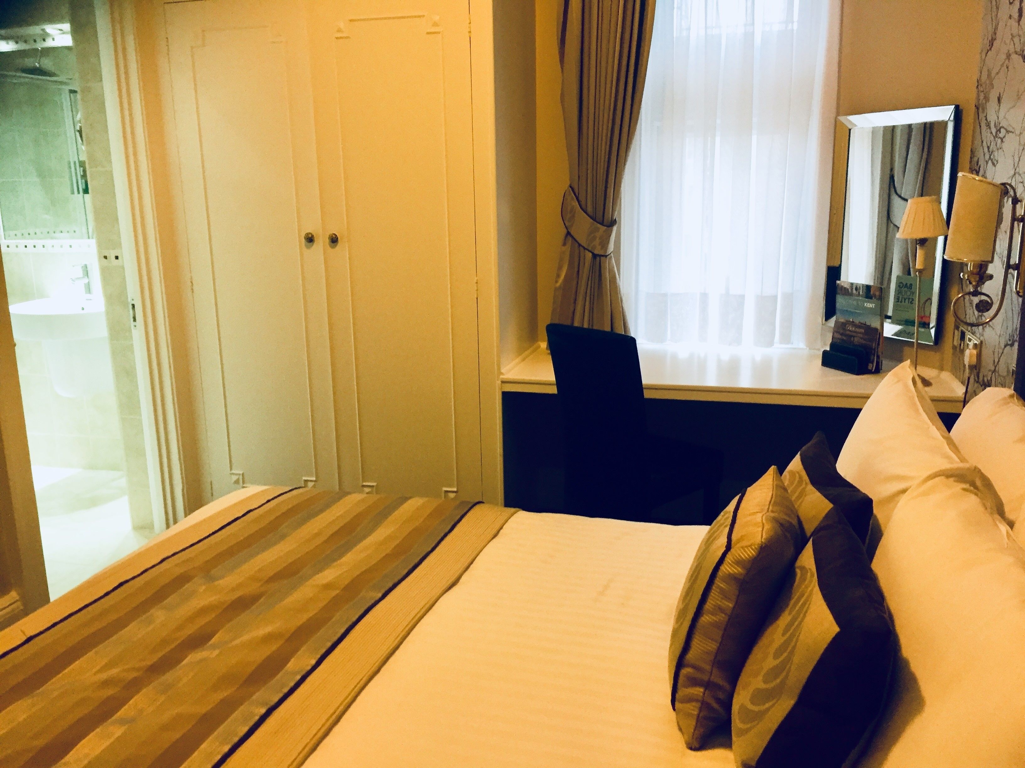 Una habitación de hotel con cama, escritorio, silla, ventana con cortinas y baño visible a través de una puerta abierta.