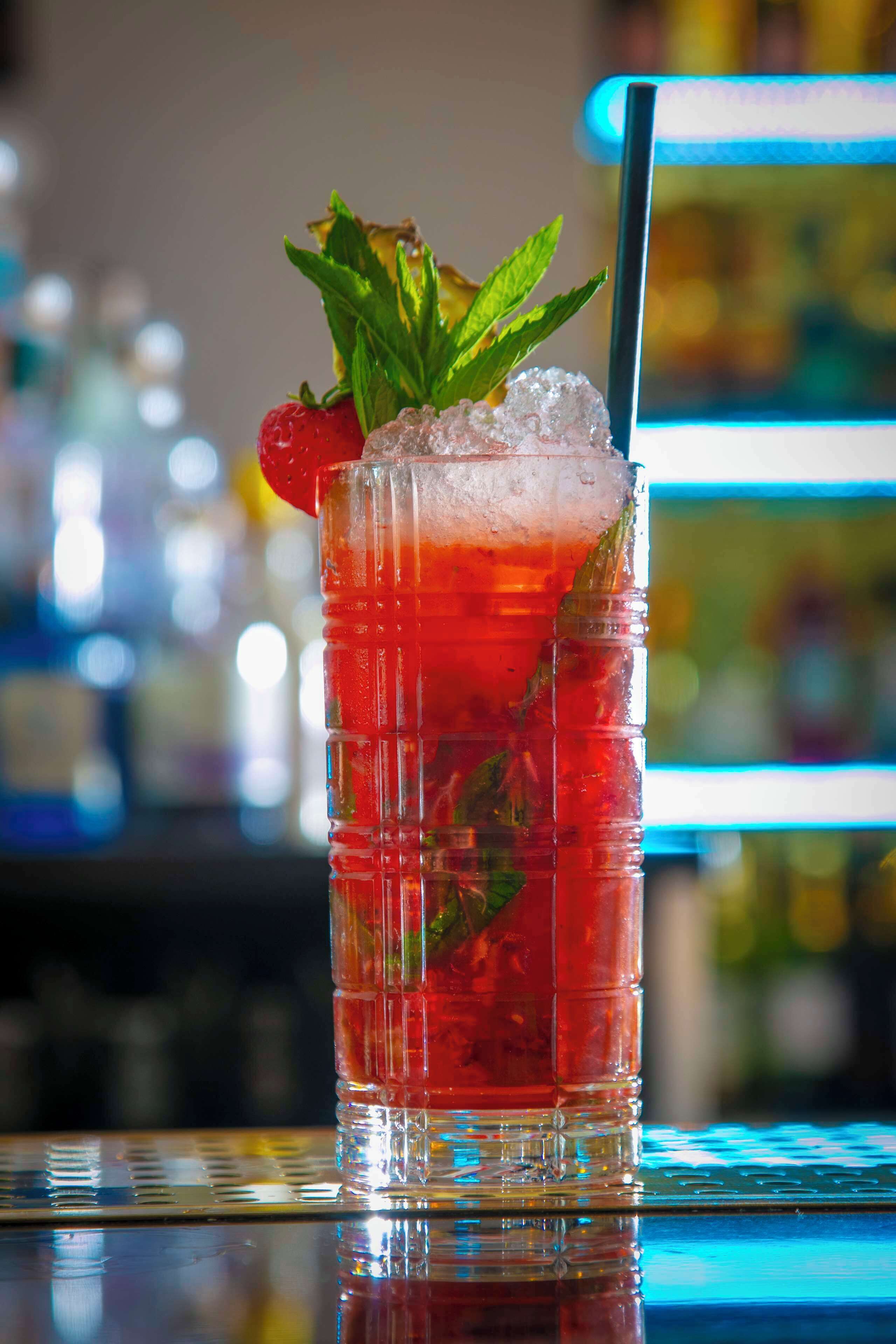 A tall glass with a red beverage, ice, a strawberry, and mint leaves.