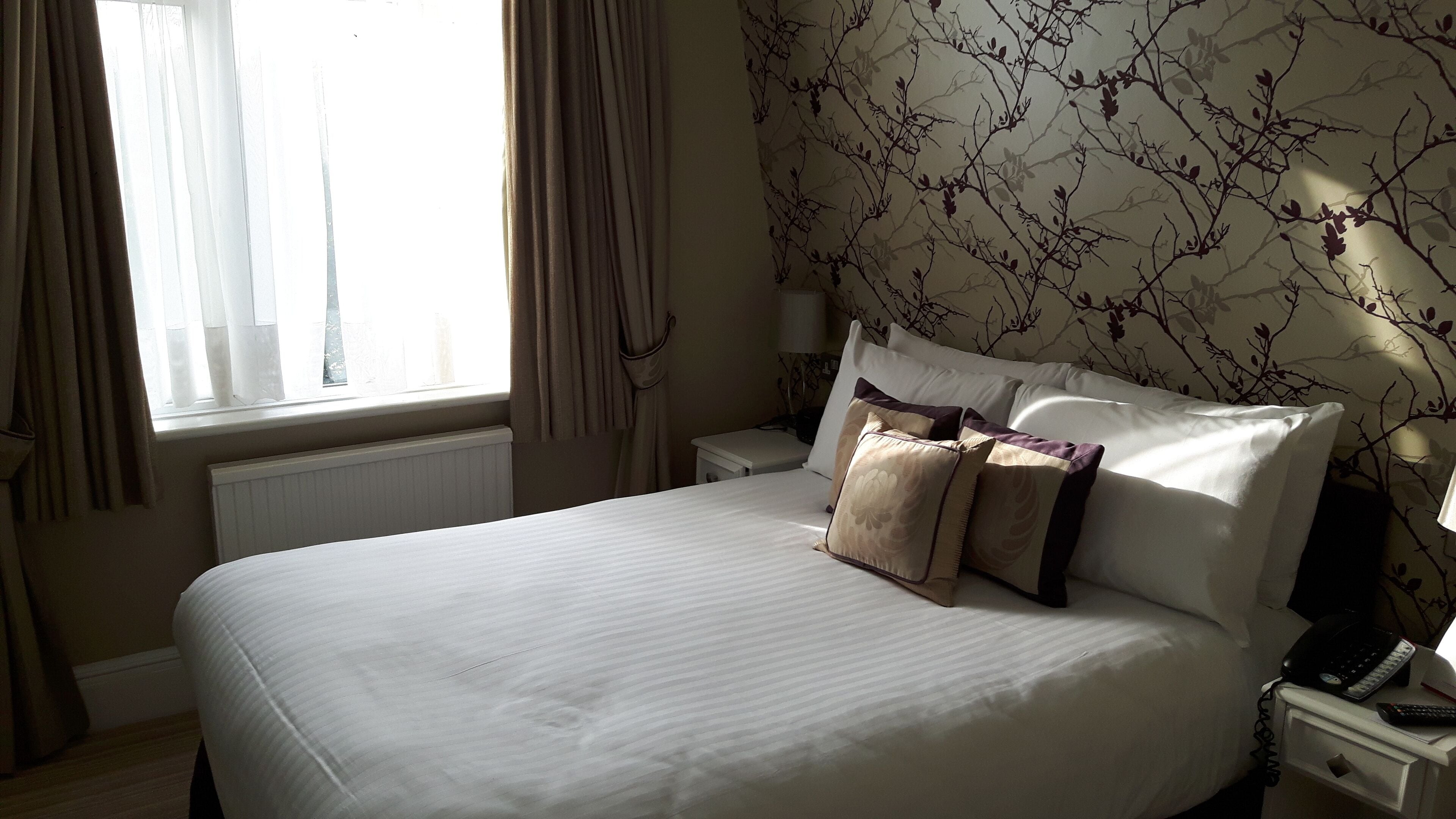 A hotel room with a large bed, a bedside table with a phone and a lamp, and a window with curtains.