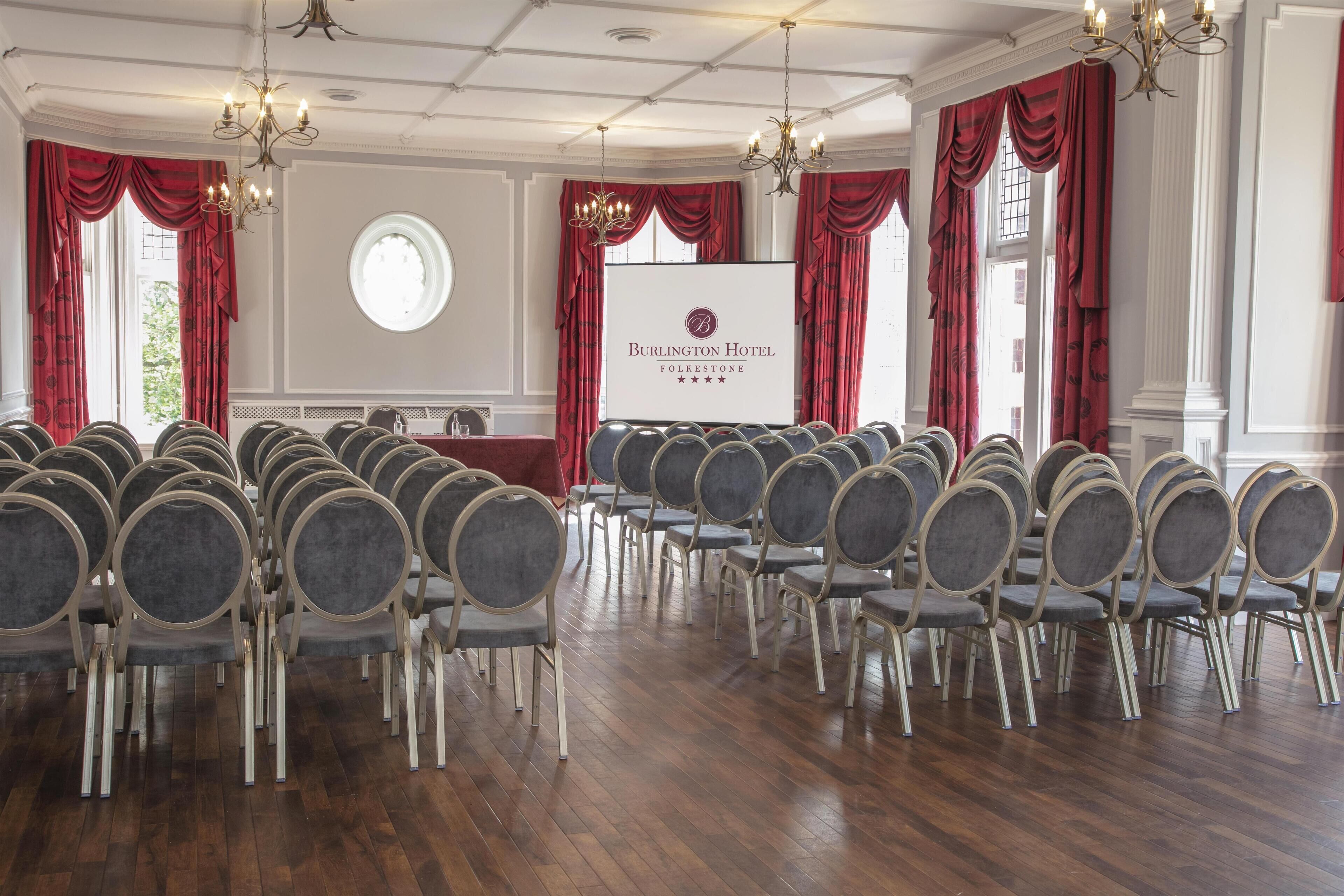 Uma sala de conferências no Hotel Burlington FolkeStone, com fileiras de cadeiras cinzas, cortinas vermelhas e lustres.