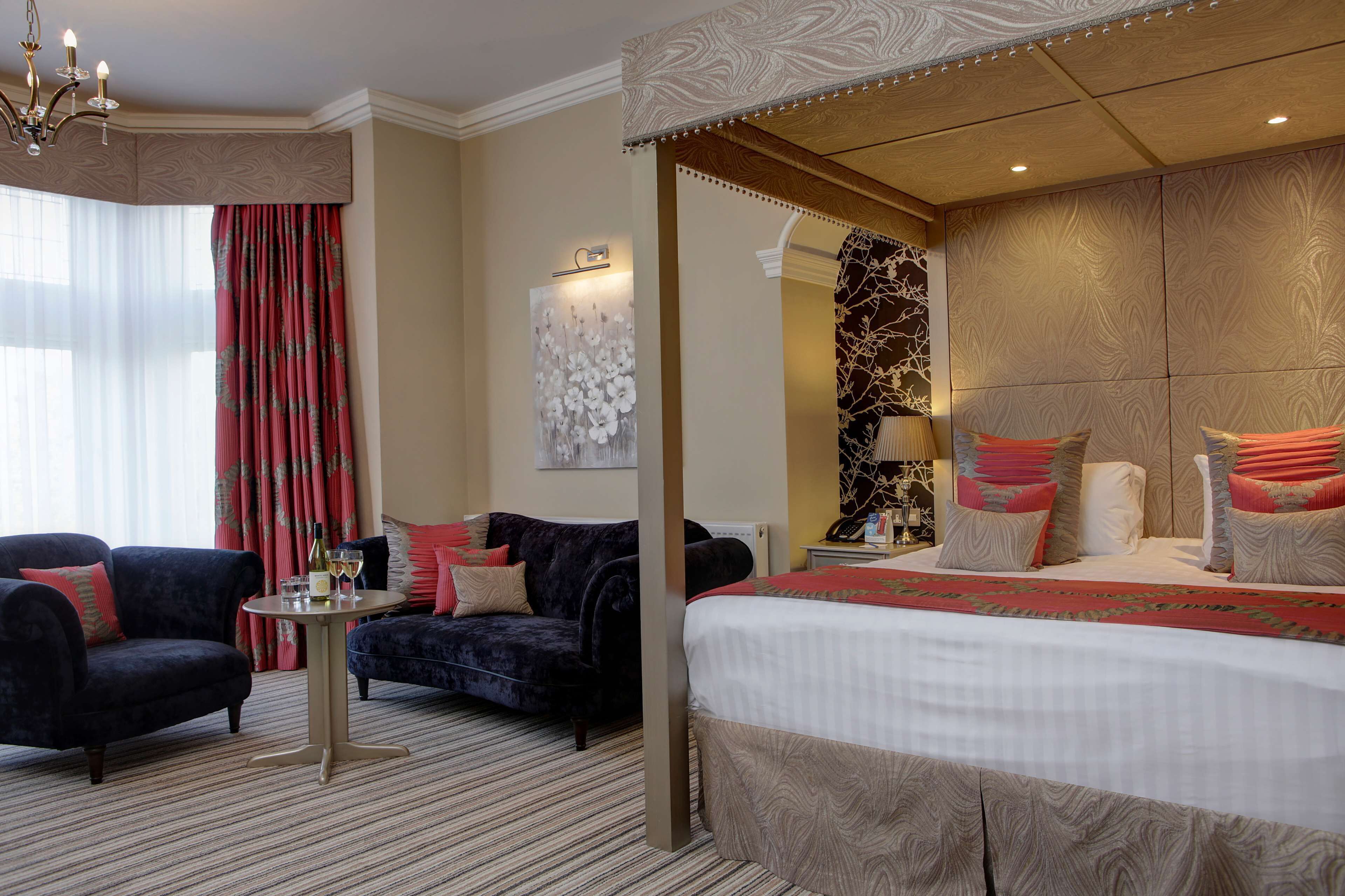 A hotel room with a large bed, two armchairs, a small table with a bottle and glasses, and a decorative wall piece.