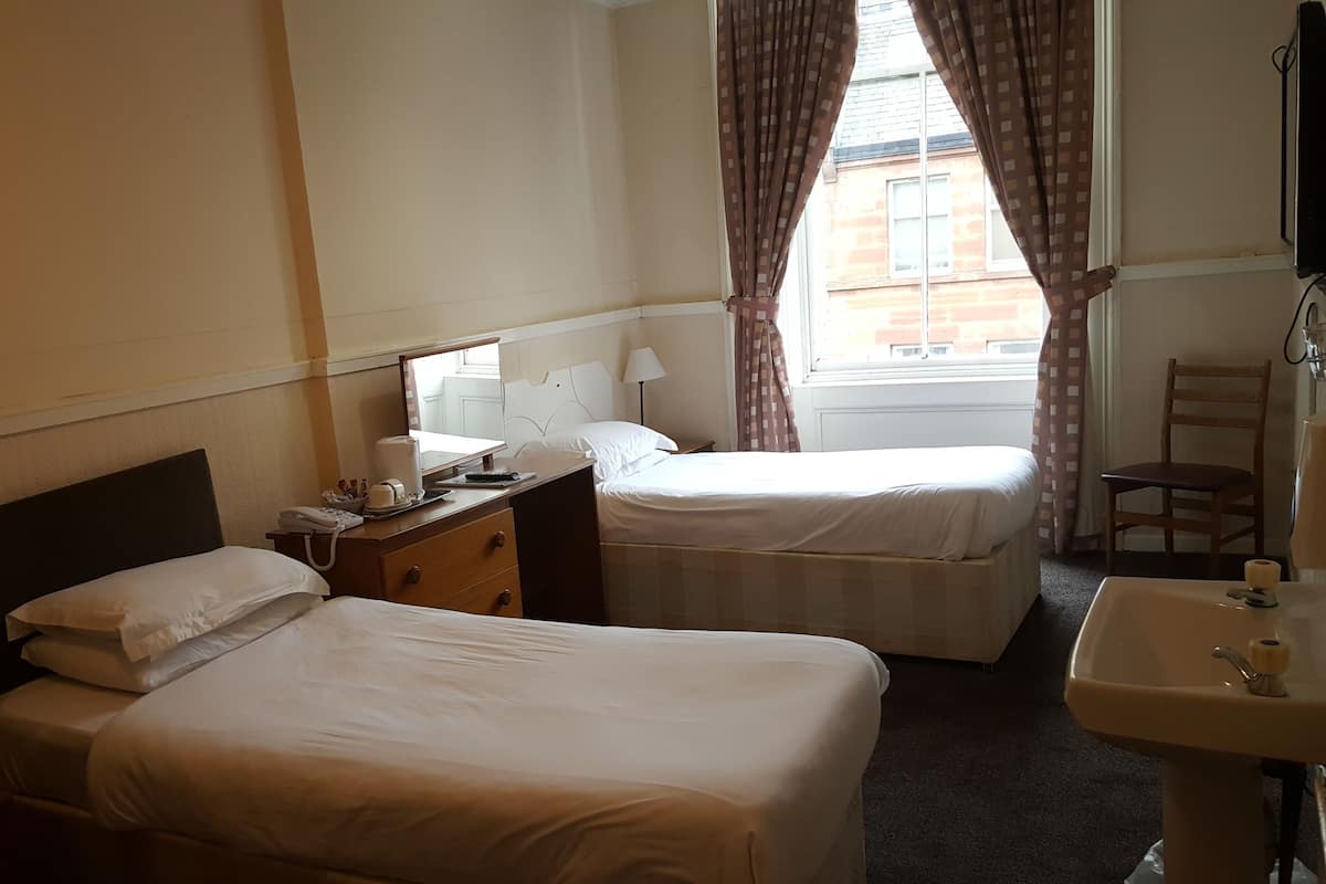 A hotel room with two single beds, a bedside table with a lamp, a small desk with a mirror, a chair, and a bathroom with a sink and mirror.