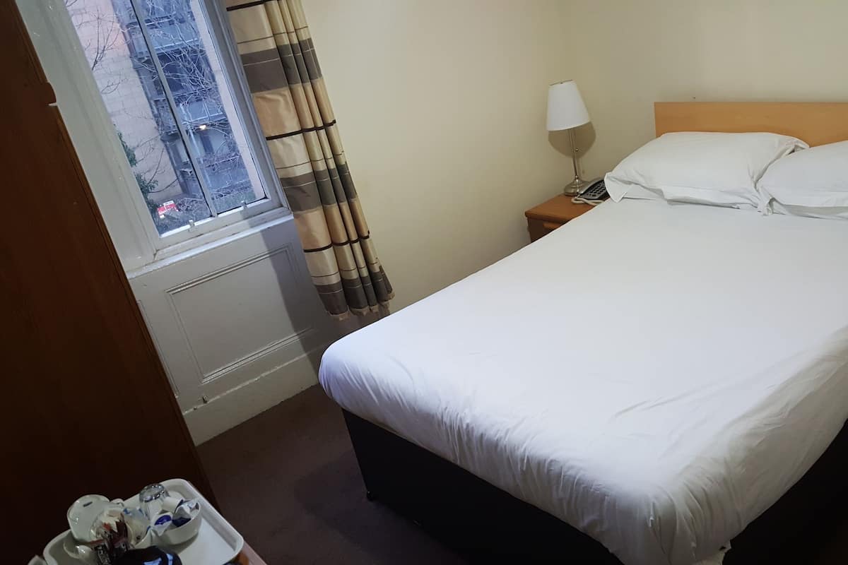 A hotel room with a bed, a nightstand, a lamp, a window with curtains, and a small table with items on it.